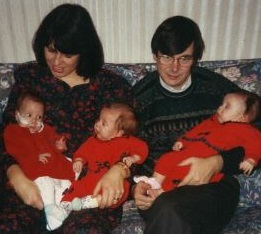 parents sit on a sofa holding three small babies 