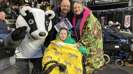 William in the stands at a football match with his parents and the team's mascot who is in a badger costume.