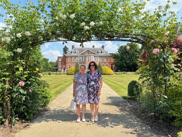 Two ladies standing side by side smiling at the camera, in the background is a mansion house