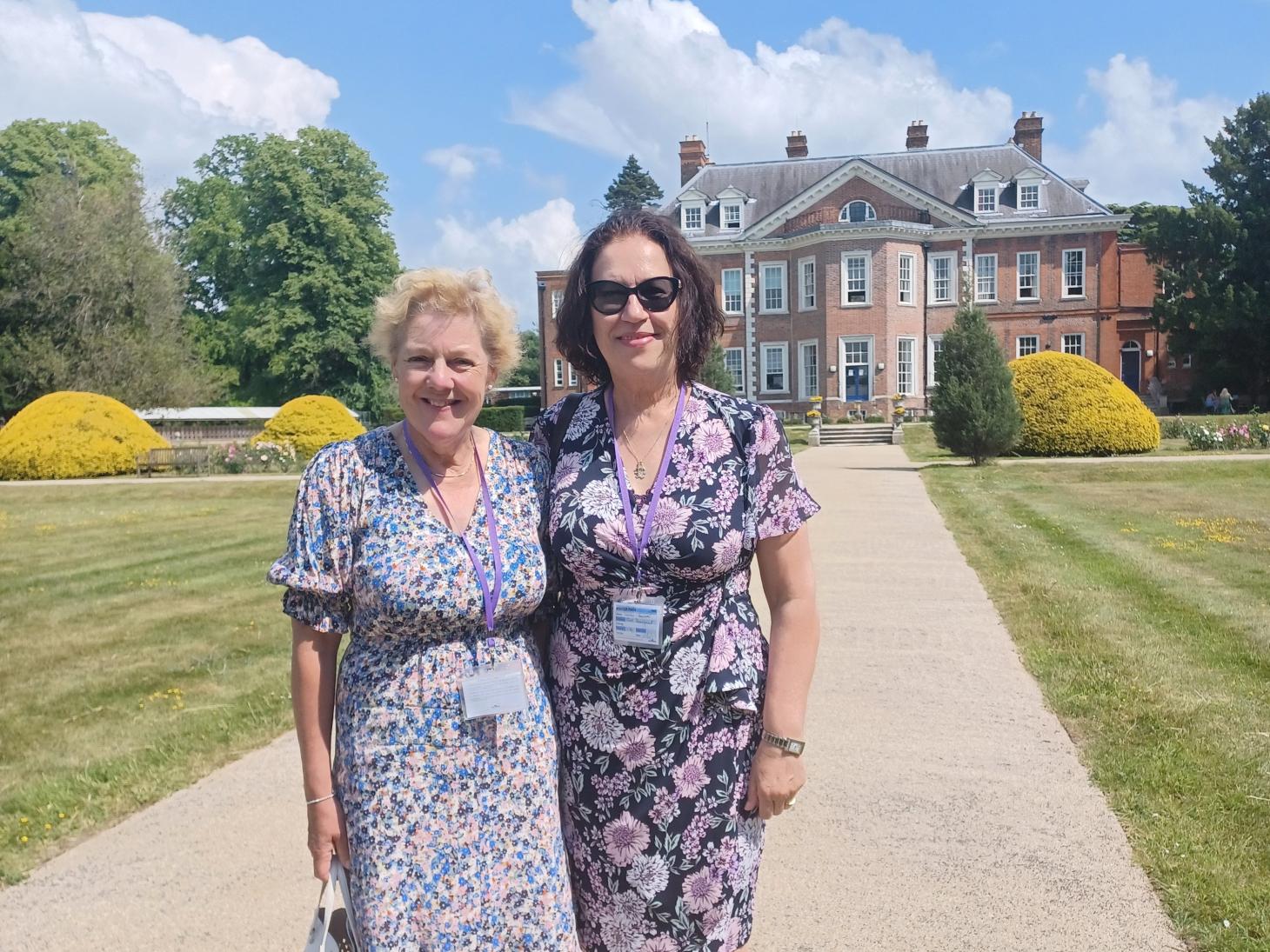 Two ladies standing side by side smiling at the camera, in the background is a mansion house