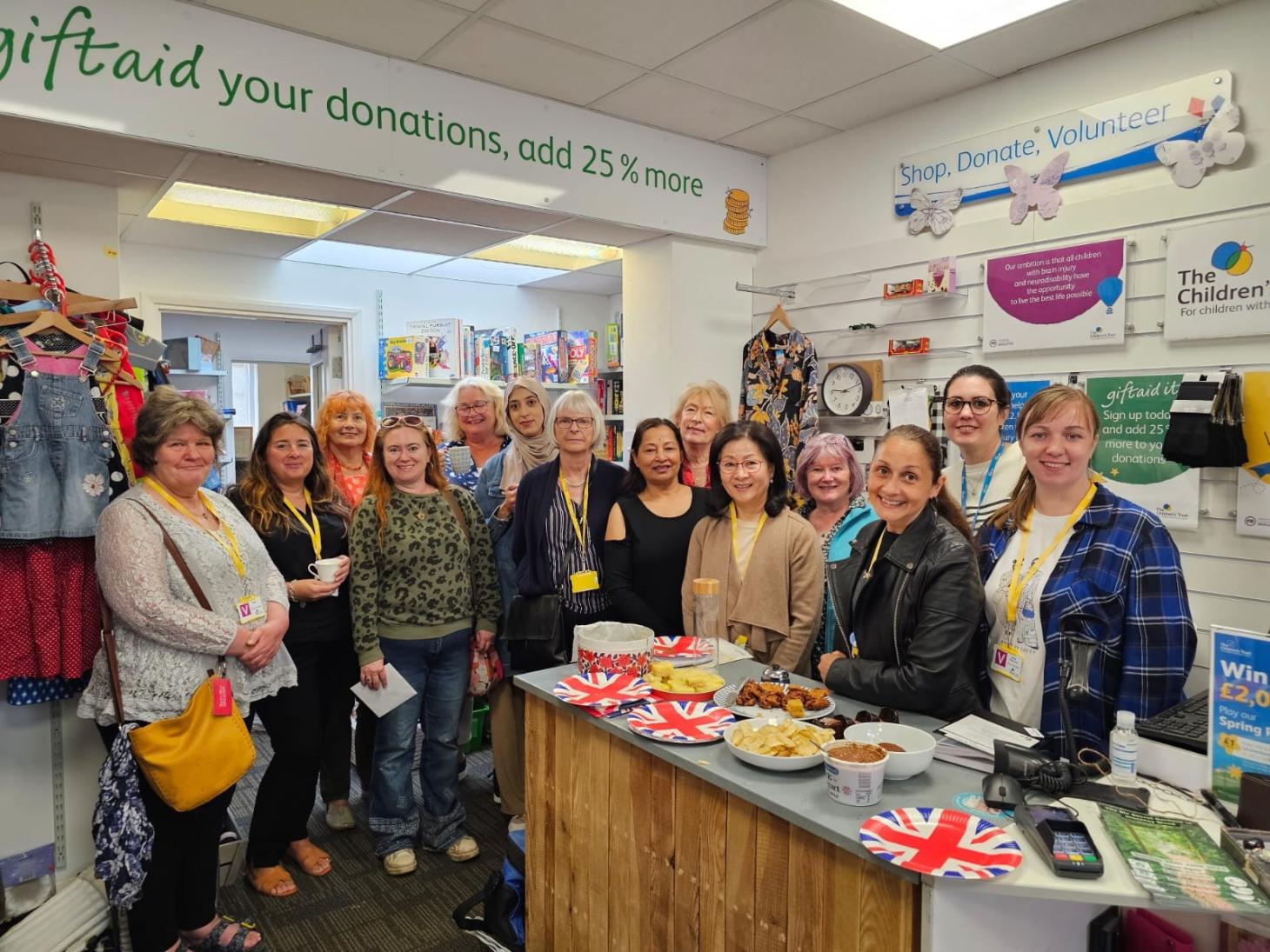 A group of volunteers stood in a charity shop. There is an afternoon tea on the table in front of them.