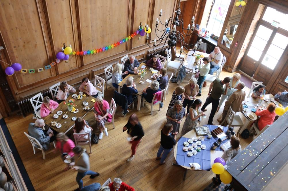 Aerial shot of a room with tables, party banners and balloons. People are standing and chatting or sitting and eating.