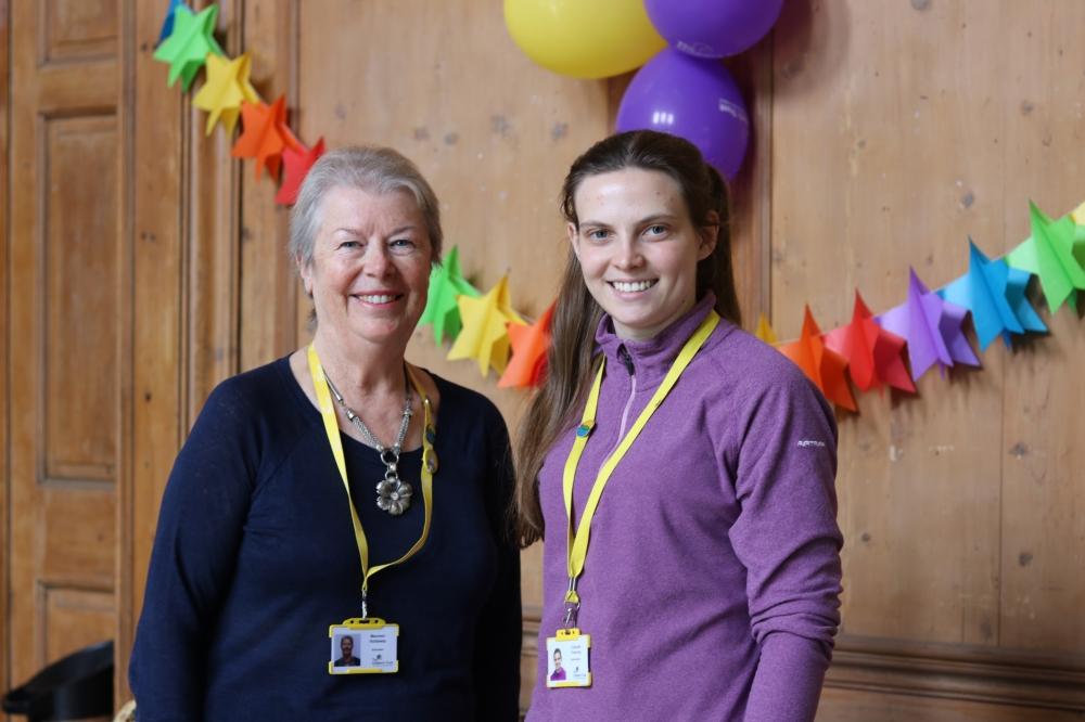 Two volunteers smiling at the camera, there are balloons and a party banner behind them