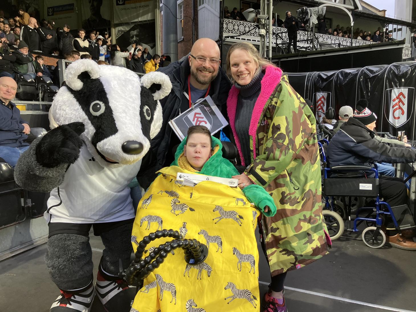 William in the stands at a football match with his parents and the team's mascot who is in a badger costume.