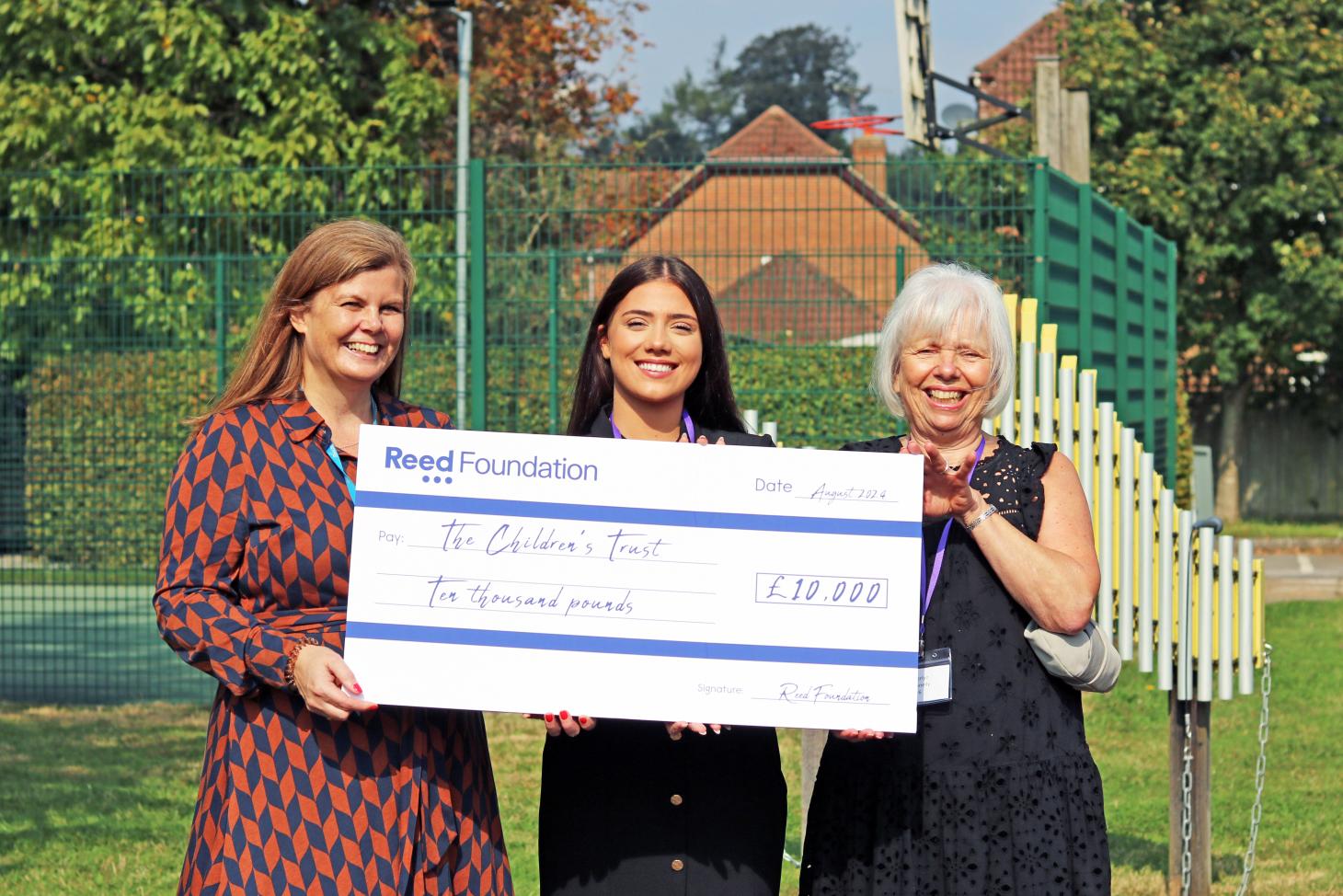 Merlyn, Freya and Michelle hold a big cheque made out to The Children's Trust for £10,000. They are smiling at the camera.