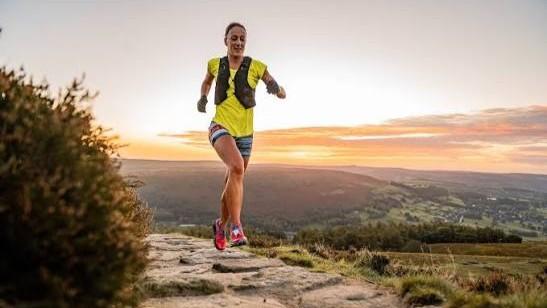 Female runner in yellow top, stripey shorts and pink trainers at sunset running along a hillside path flanked with lush green grass and a picturesque orange sky