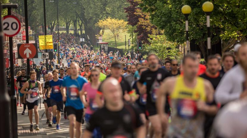 blurred runners filling the street next to a zebra crossing and a 20mph sign amongst lush green trees